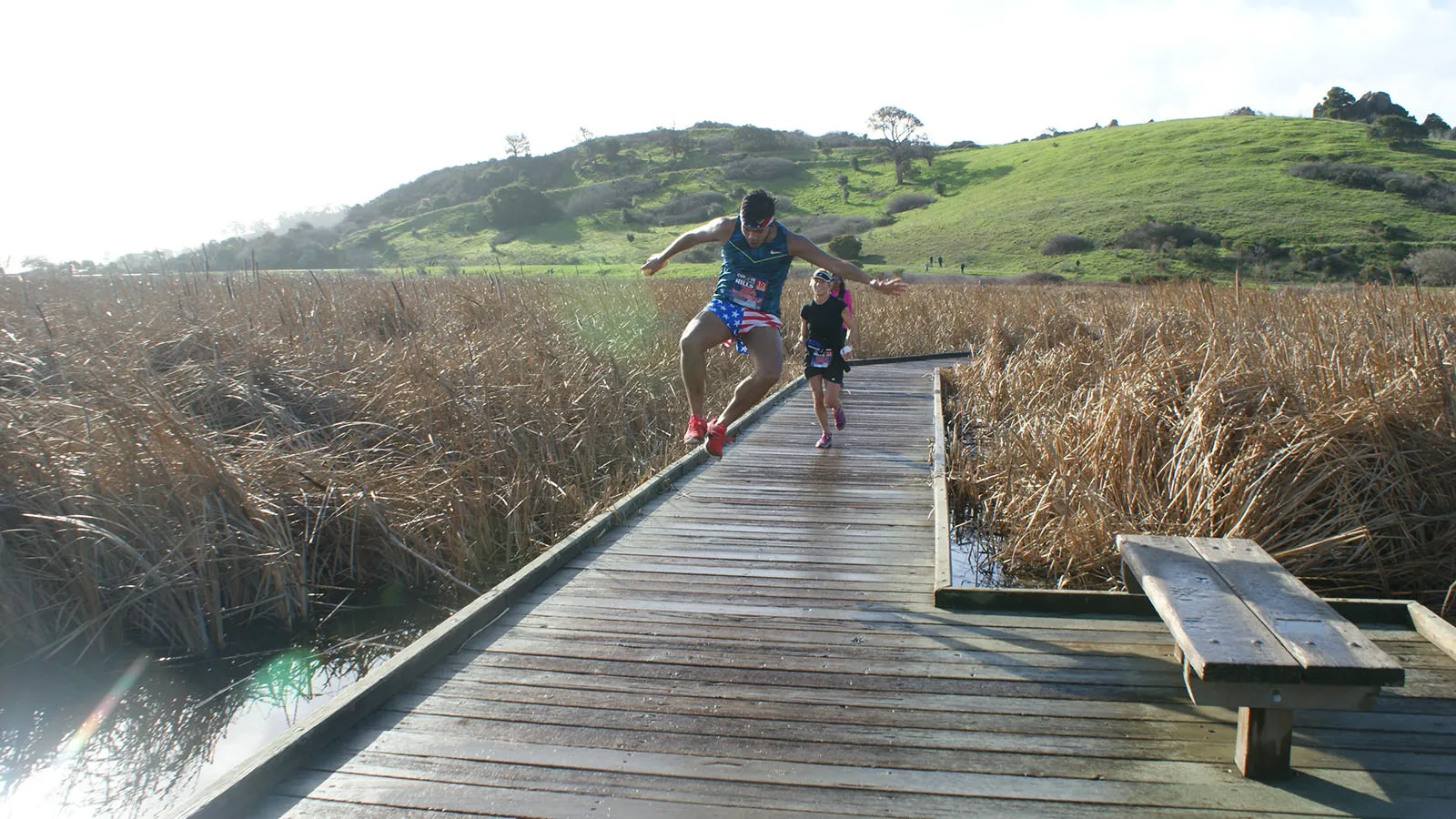 A runner jumps in the air on a wooden boardwalk, with another runner behind him and a grassy hill in the background.
