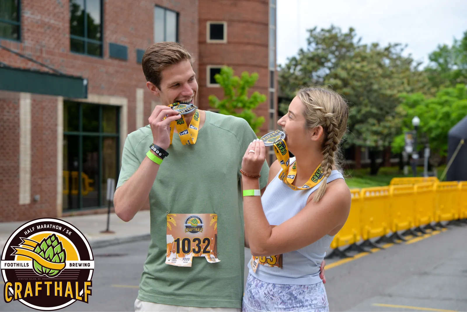 A man and a woman smile at each other while holding up and admiring their race medals, with bib numbers visible on their shirts.