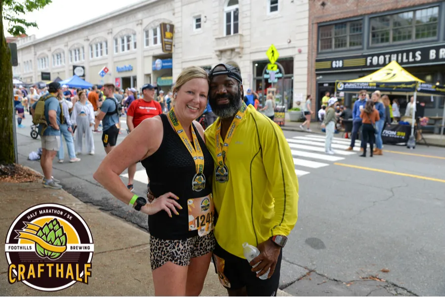 A smiling couple, both wearing race medals, pose for a photo on a street with other runners and tents in the background.