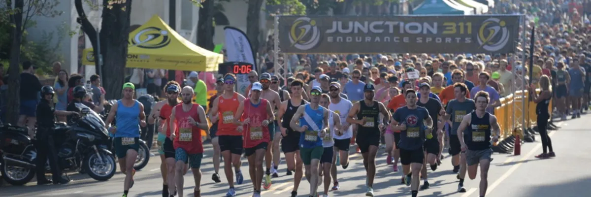 A large group of runners starting a race, with a banner overhead that says 'JUNCTION 311 ENDURANCE SPORTS'.