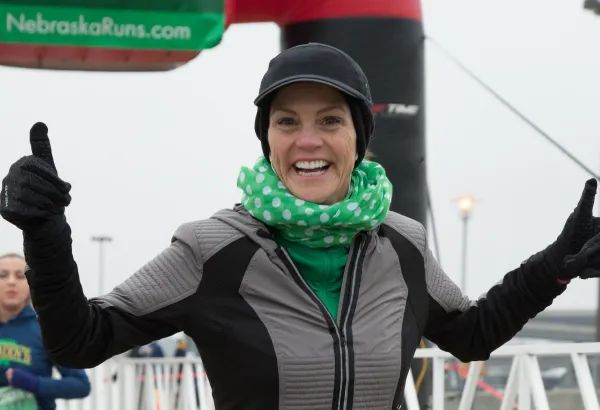 A woman smiles and gives a thumbs up at the finish line of a race, wearing a green polka-dot scarf.