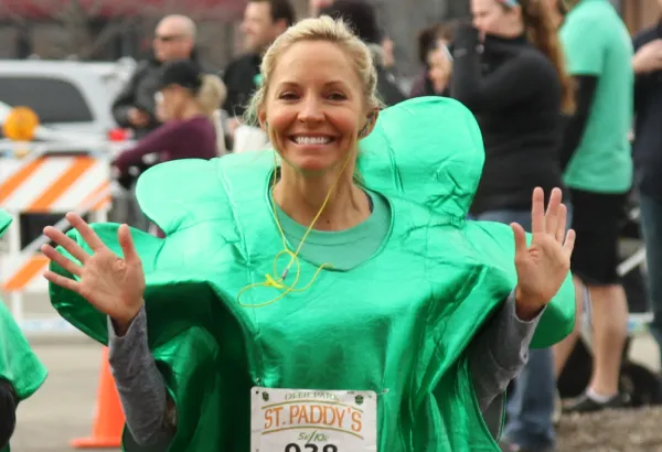 A woman in a green shamrock costume smiles and waves at the camera during the St. Paddy's 7K race.