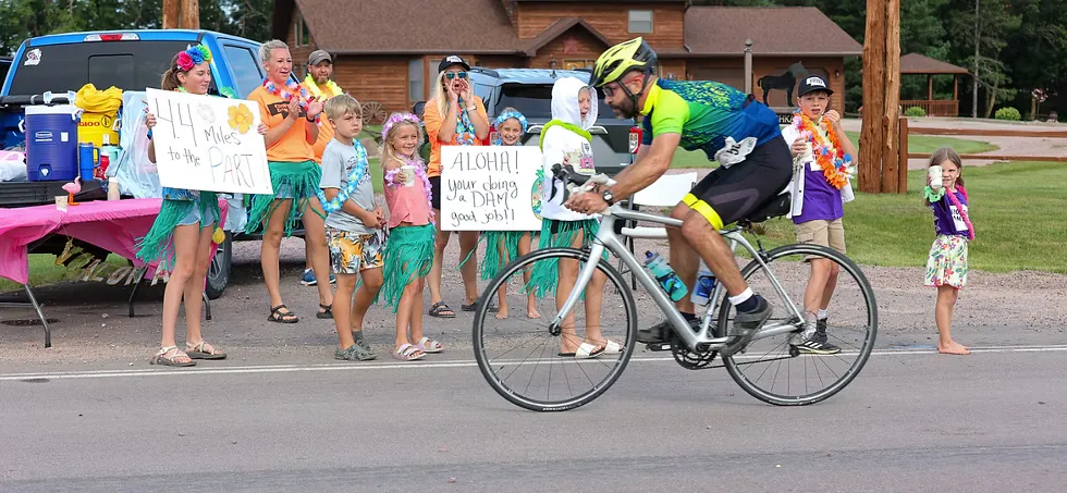 A cyclist passes a group of people holding signs and cheering, some wearing leis and grass skirts.