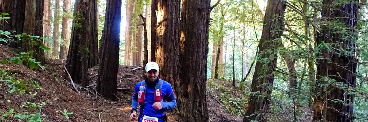 A smiling male runner wearing a blue jacket and a white cap runs on a wooded trail, with large trees surrounding him.