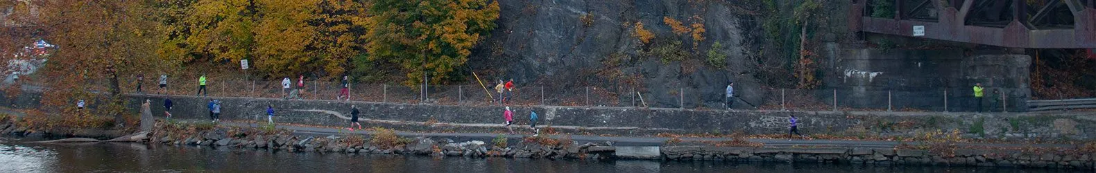 Runners on a path alongside a river with autumn foliage and a bridge in the background.
