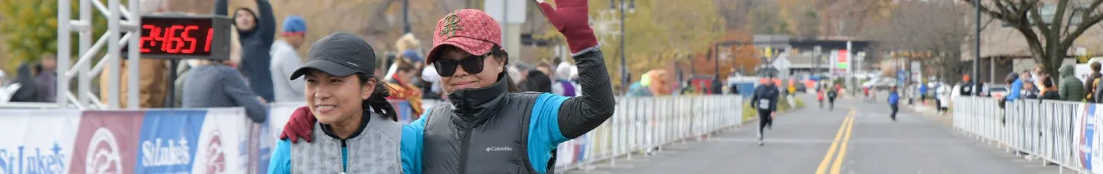 Two women smiling and waving at the start line of a race, with a digital clock showing the time in the background.