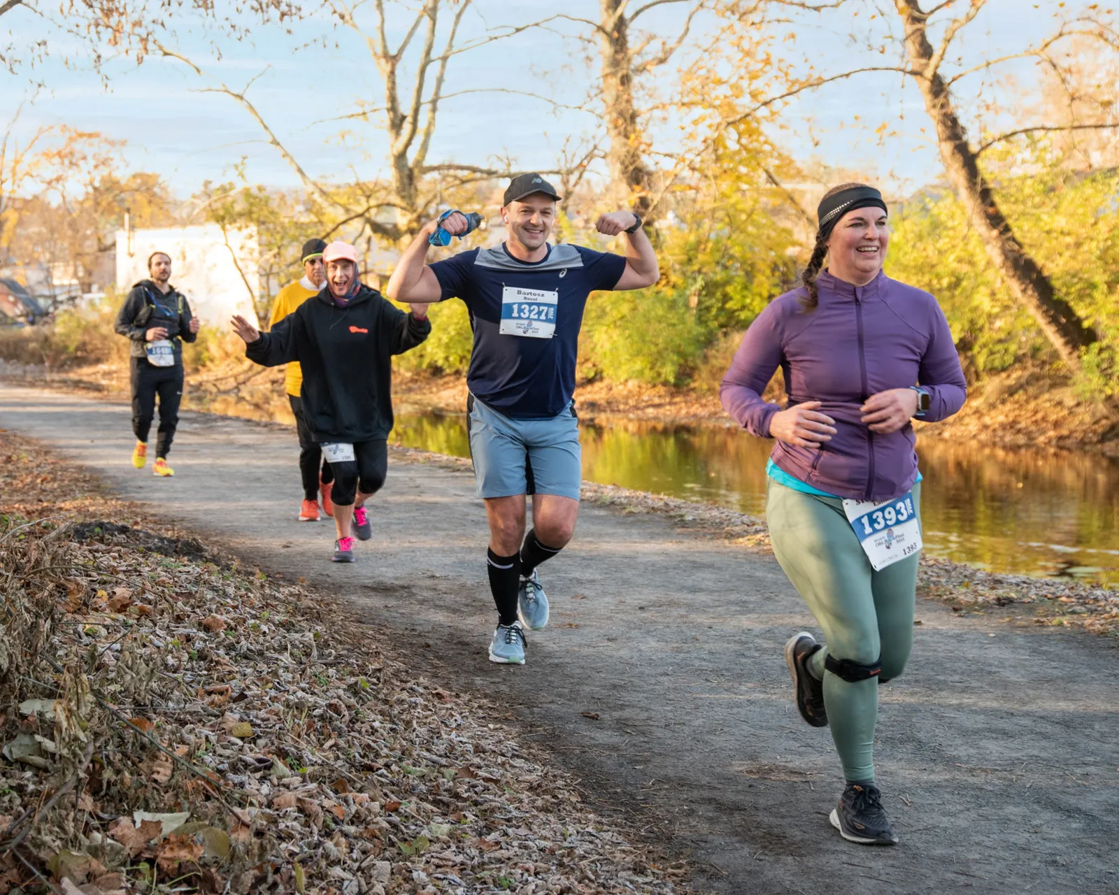 Runners on a path next to a body of water, with one runner flexing his bicep and another smiling.