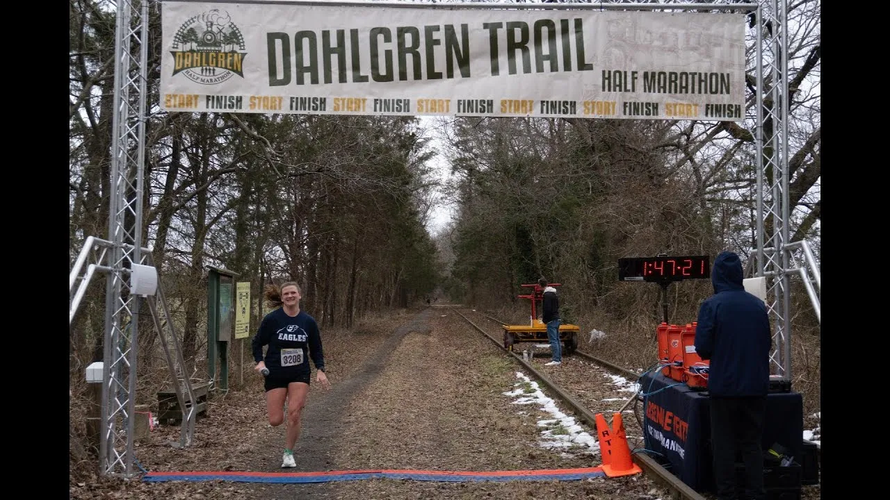 A female runner crosses the finish line of the Dahlgren Trail Half Marathon, with a large banner overhead and a digital clock displaying the time.