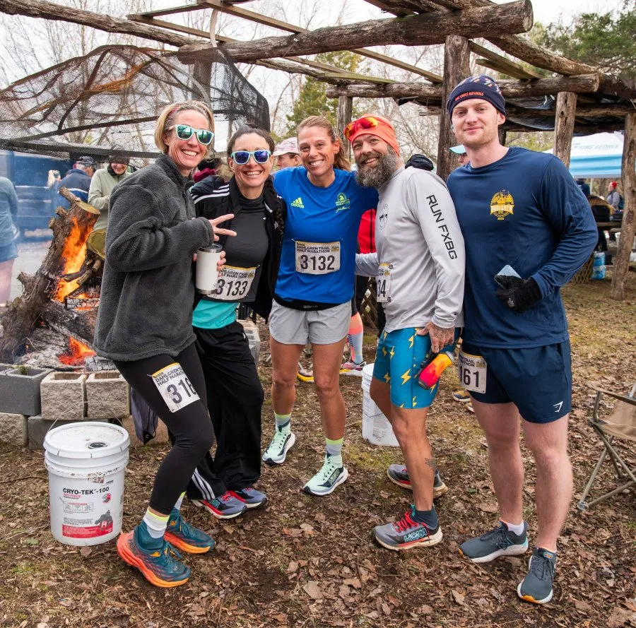 A group of five runners smile and pose for a photo in front of a bonfire and wooden structure at the Dahlgren Trail Half Marathon.