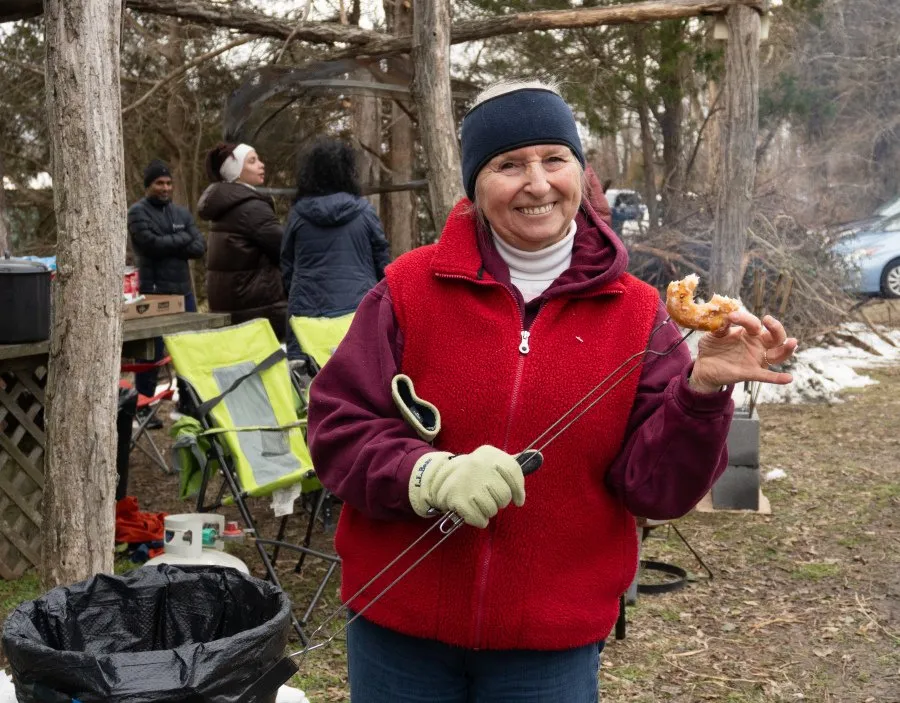 A smiling woman in a red vest holds a donut on a skewer, with other people and a wooded background.