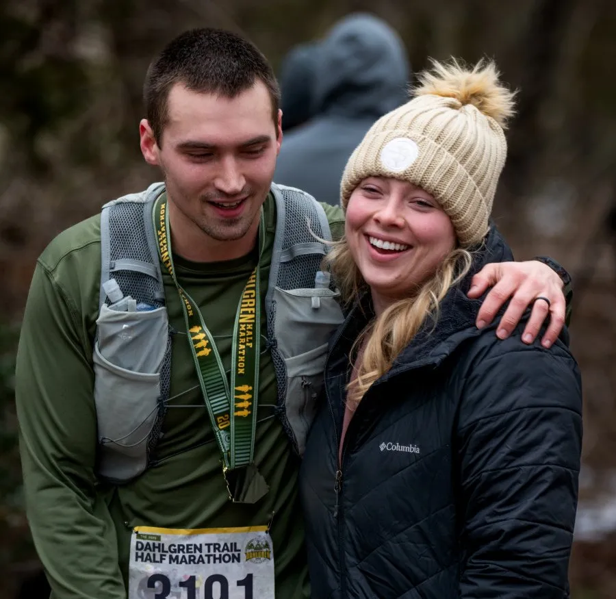 A smiling couple, a man and a woman, wearing race medals and bibs, with the woman's arm around the man's shoulder.