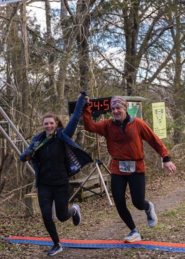 A woman and a man celebrate as they cross the finish line of a trail race, with a digital timer in the background.