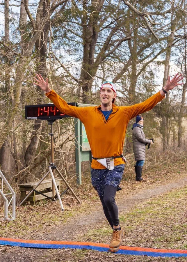 A male runner in an orange long-sleeved shirt and blue shorts crosses the finish line with his arms raised in triumph, a digital timer showing 1:44 in the background.