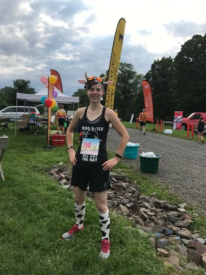 A runner wearing a cow-themed costume, including a headband with horns and cow-print socks, stands with hands on hips at a race event.