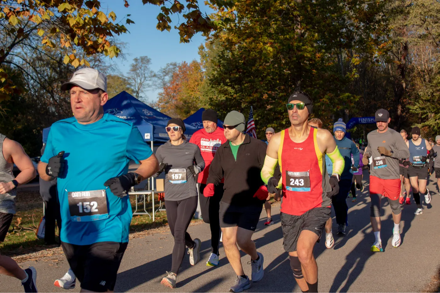 Runners in a race, with bib numbers visible, running on a paved path lined with trees.