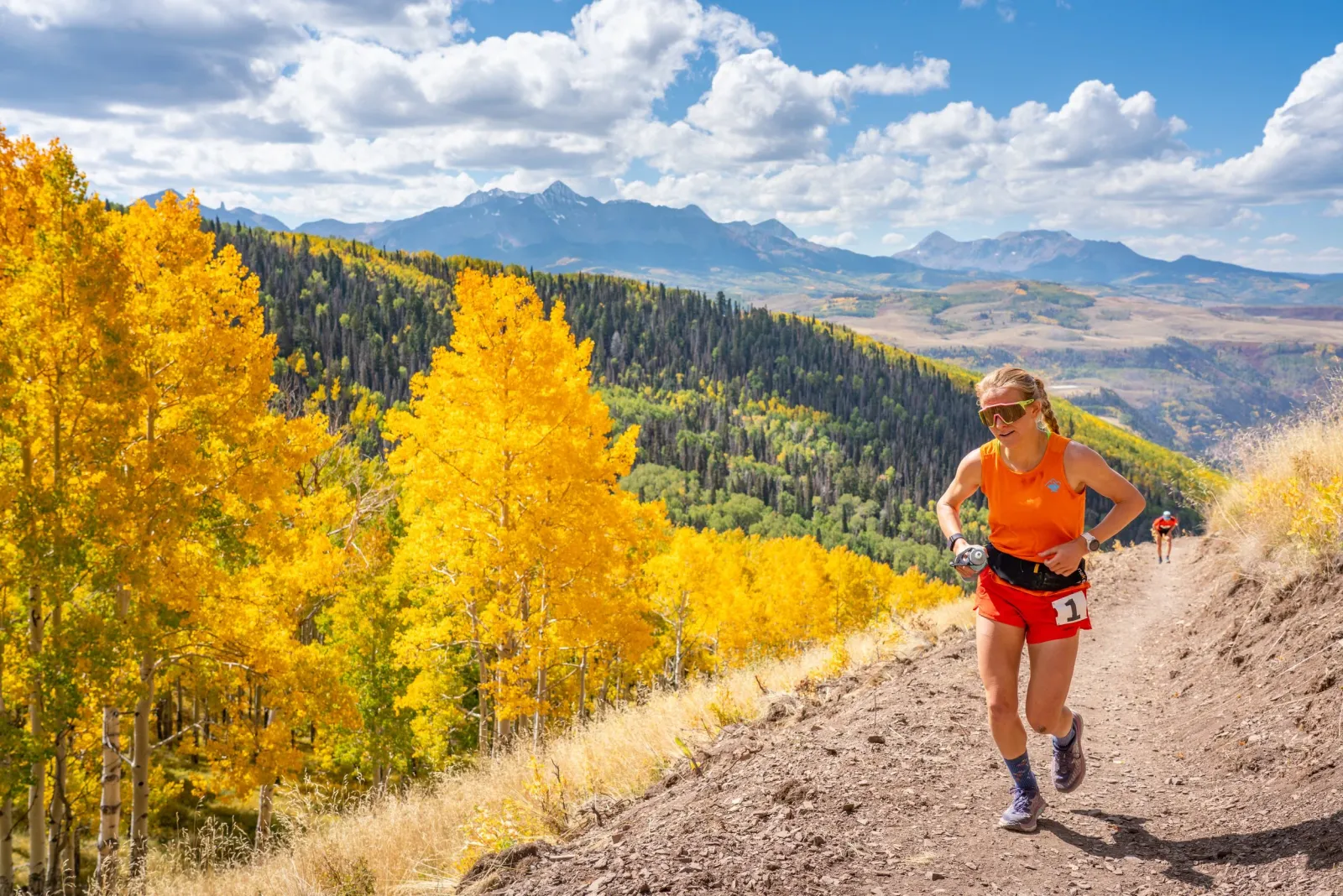 A female runner with blonde hair in a ponytail, wearing an orange tank top and red shorts, runs on a dirt trail with a scenic mountain and autumn foliage background.