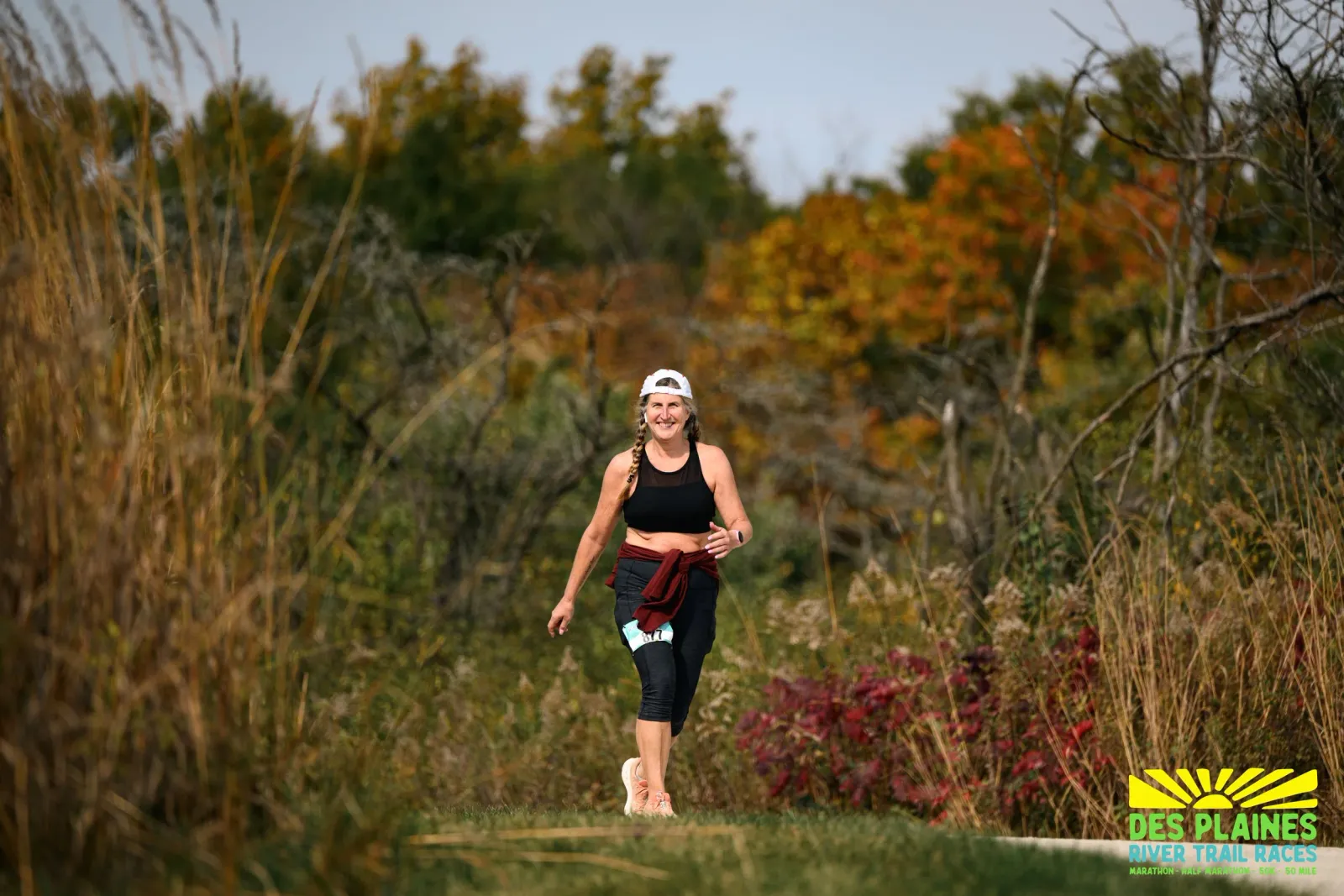 A female runner smiles as she runs on a trail with autumn foliage in the background.
