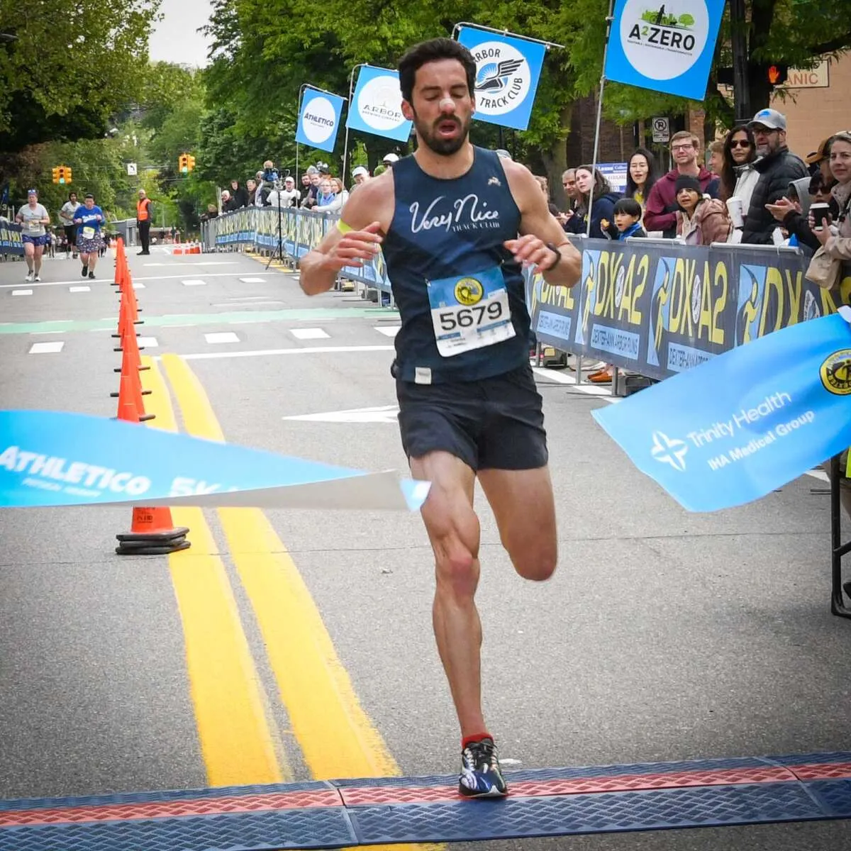 A male runner crosses the finish line of a road race, breaking through a blue ribbon.
