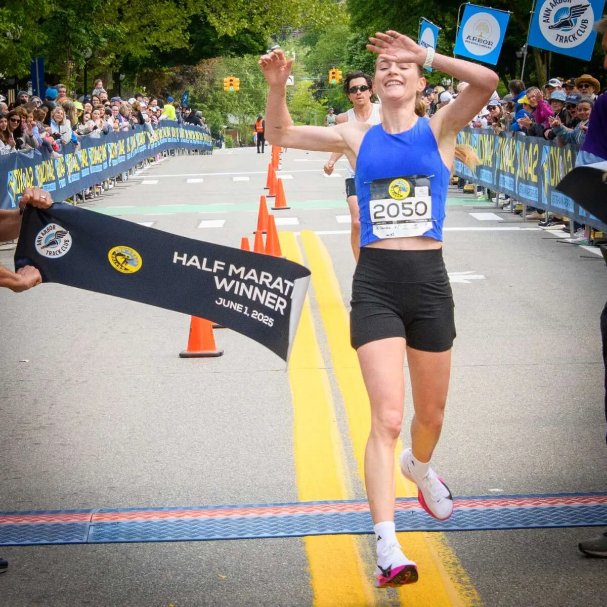 Female runner crossing the finish line with arms raised in celebration, holding a banner that says 'Half Marathon Winner'.