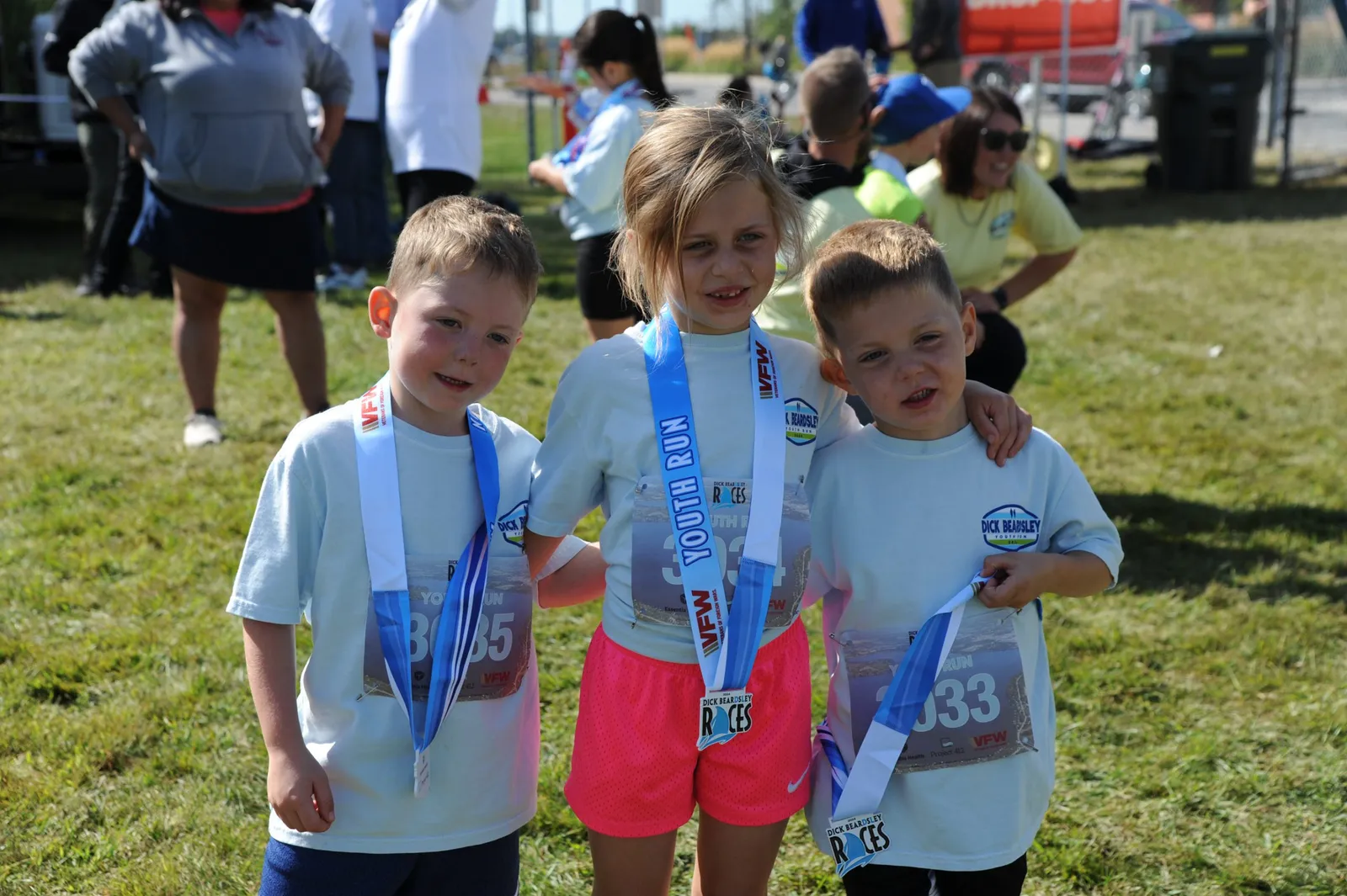 Three young children, two boys and a girl, wearing race medals and t-shirts, smiling at the camera.
