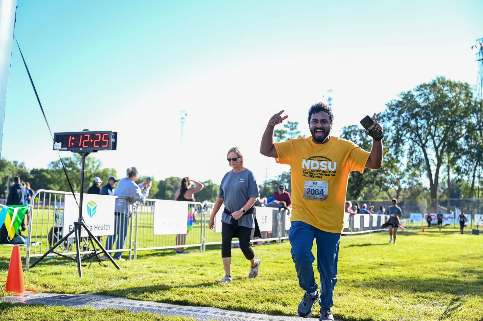 A runner in a yellow t-shirt with a bib number 2064 crosses the finish line, smiling and giving a peace sign with one hand while holding a phone in the other. A digital clock showing 1:12:25 is visible in the background.
