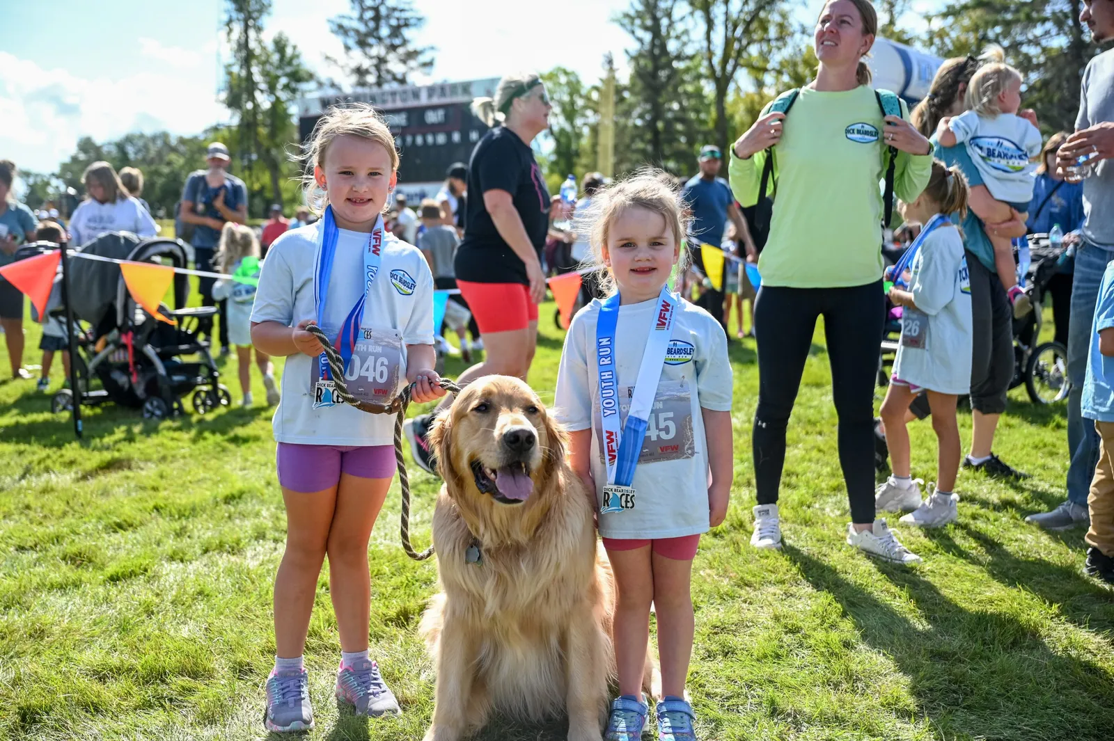 Two young girls wearing race medals and t-shirts stand with a golden retriever dog on a grassy field, with other participants and a race banner in the background.
