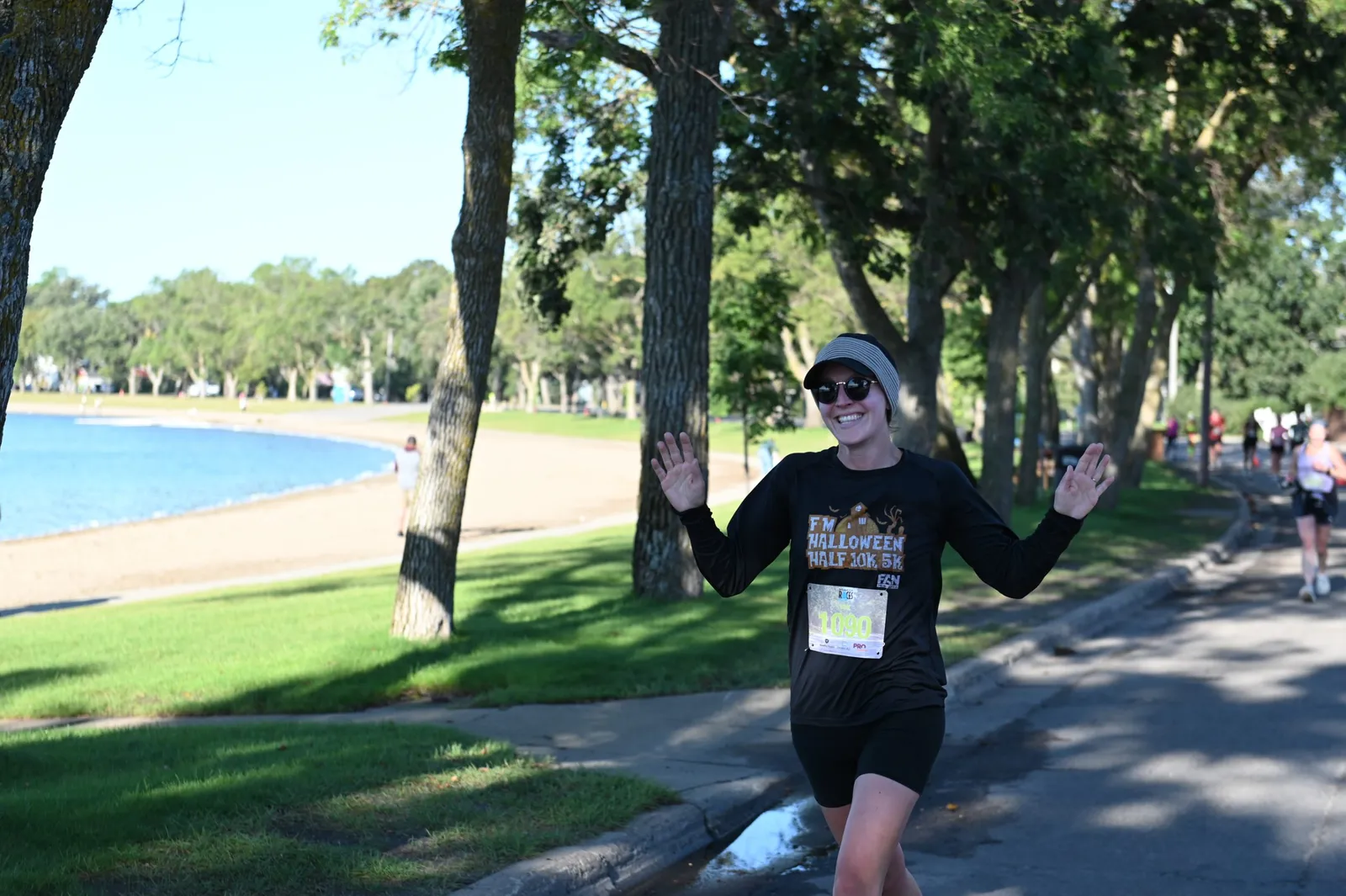 A smiling female runner wearing sunglasses and a black long-sleeved shirt with a bib number waves to the camera while running on a road next to a body of water.