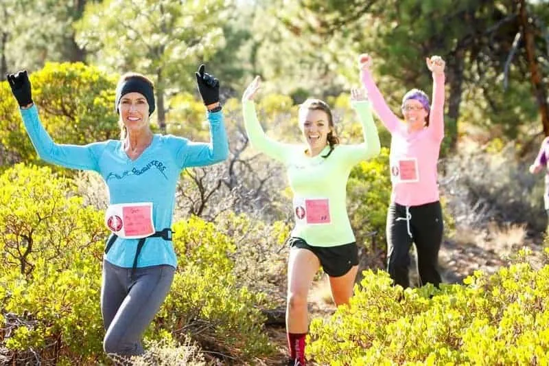 Three women running a trail race, with two in the foreground raising their arms in celebration.