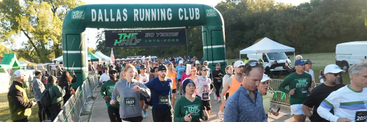 A large group of runners are starting a race under a green inflatable archway that says "Dallas Running Club" and "The Half".