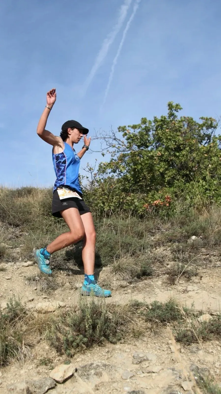 A female runner with her arms raised in excitement, running uphill on a dusty trail under a clear blue sky.
