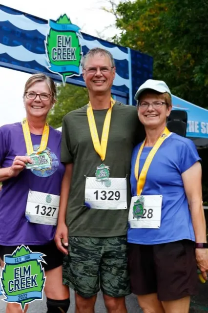 Three smiling runners, two women and one man, pose for a photo wearing medals and race bibs in front of an Elm Creek race banner.
