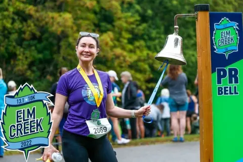 A smiling female runner wearing a medal and bib number rings a large silver bell at the Elm Creek race event.