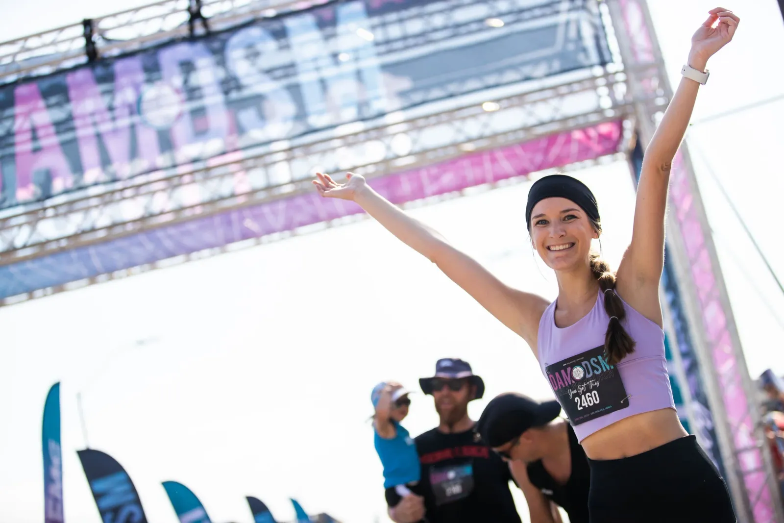 A smiling female runner with her arms raised in celebration at the finish line of the DAM to DSM race, with the race banner visible in the background.