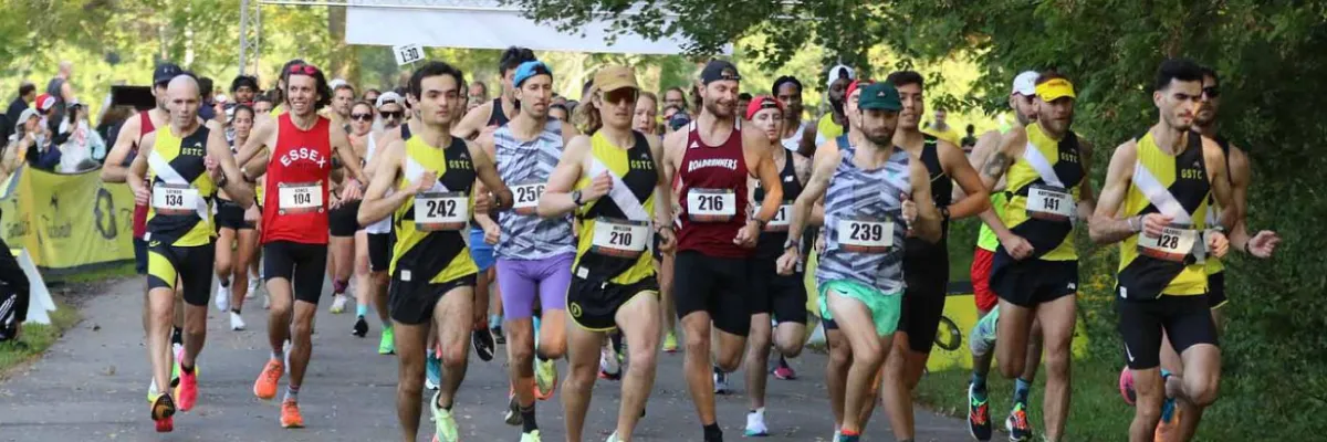 A group of runners in a race, with bib numbers visible, running on a paved path lined with trees.