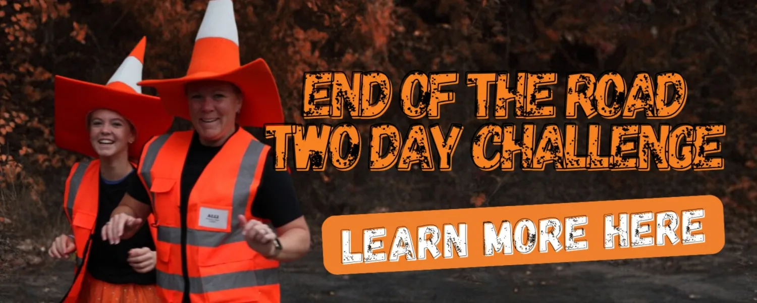 Two people wearing traffic cone hats and orange safety vests, smiling and appearing to run or dance, with the text "END OF THE ROAD TWO DAY CHALLENGE" and "LEARN MORE HERE" overlaid on the image.