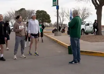 Runners gather at the start line, some looking towards an official speaking.