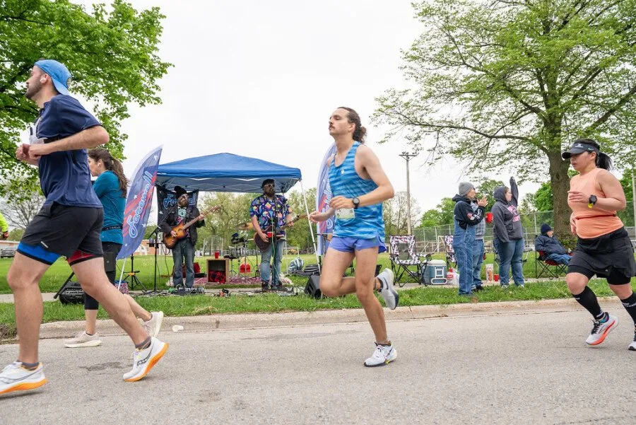 Runners pass a live band performing at the side of the road during a race, with spectators watching.