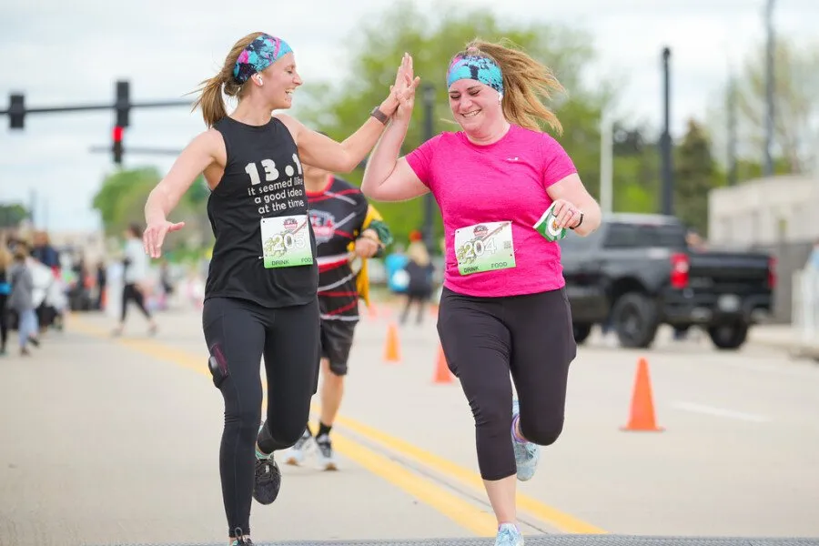 Two female runners high-fiving during a race, one wearing a black tank top with '13.1 it seemed like a good idea at the time' and the other in a pink t-shirt.