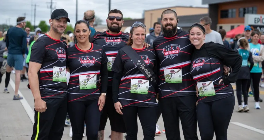 A group of six people, all wearing matching black t-shirts with 'EPIC MARATHON' logos, pose for a photo at the start of a race. One person wears a 'Birthday Girl' sash.