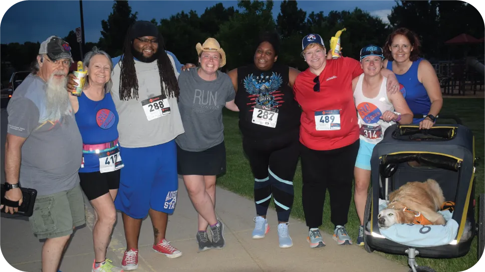 A group of eight people, including a dog in a stroller, pose for a photo at a race event. Several people are wearing race bibs.