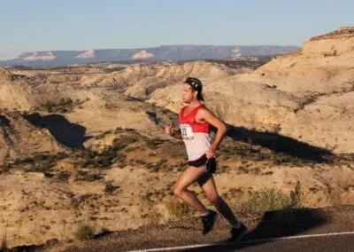 A male runner in a red and white singlet runs on a road with a scenic desert canyon landscape in the background.