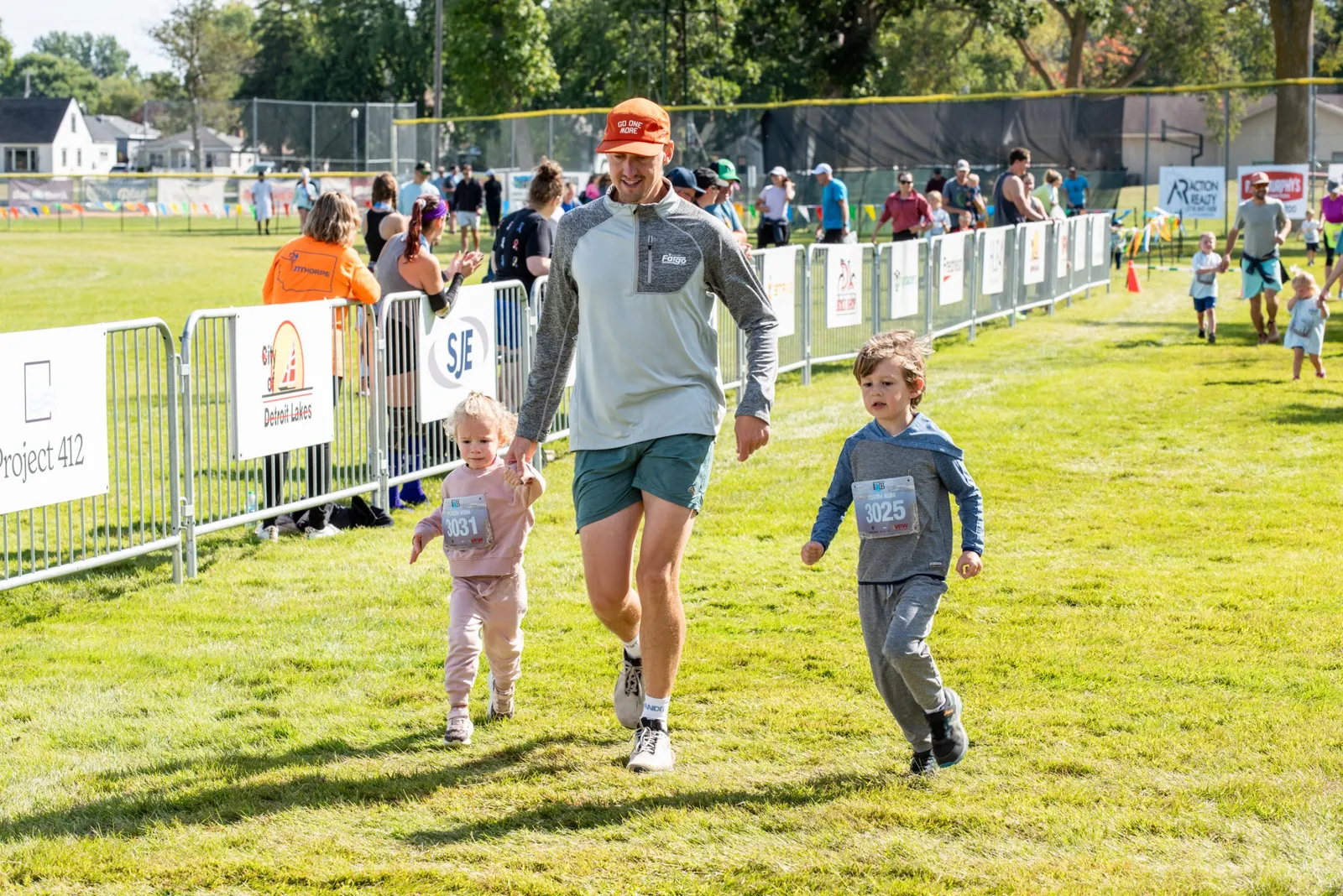 A man runs alongside two children, one boy and one girl, on a grassy field during a race.