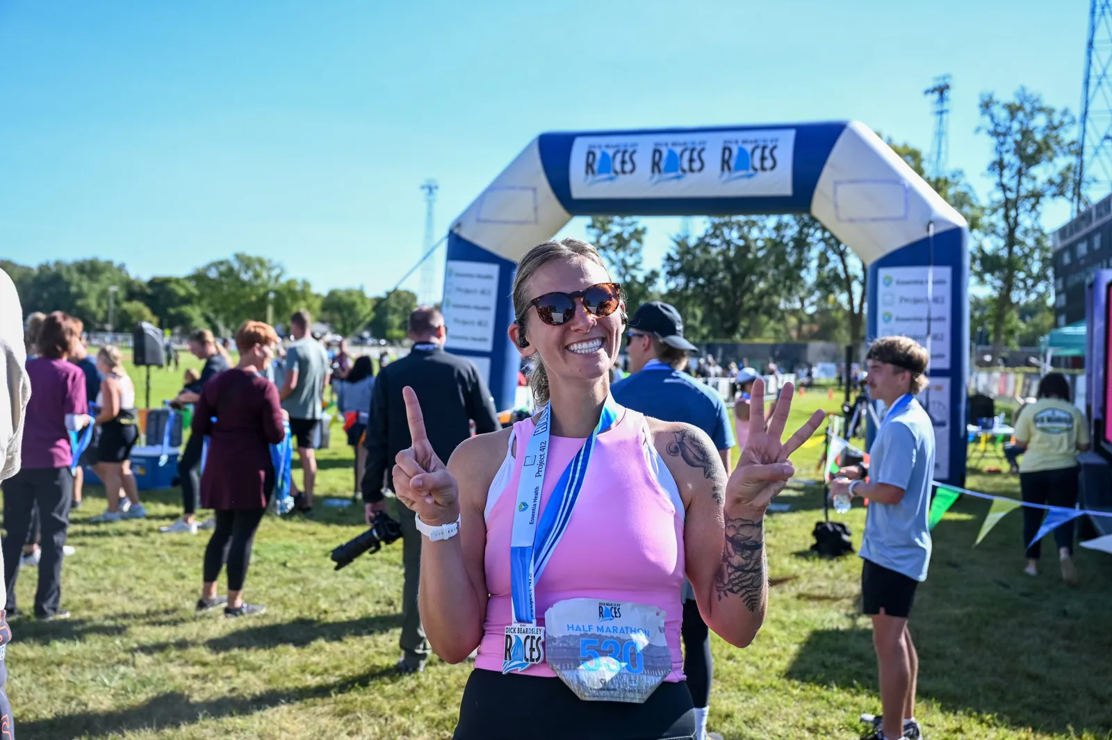 A female runner wearing a pink tank top and sunglasses smiles at the camera, holding up two fingers and wearing a race medal and bib that says 'Half Marathon 530'.