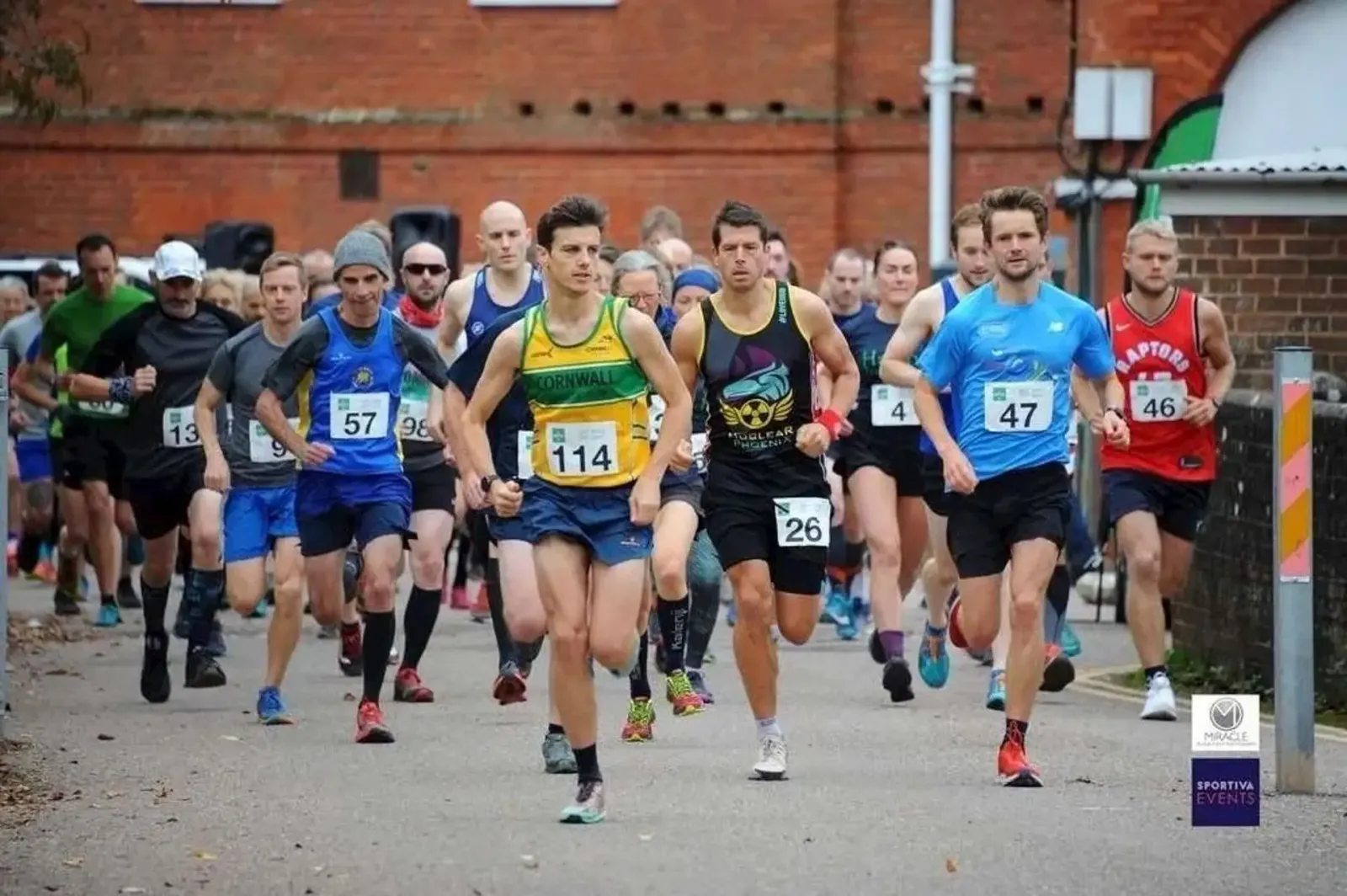 A group of male runners in racing bibs are running forward at the start of a race.