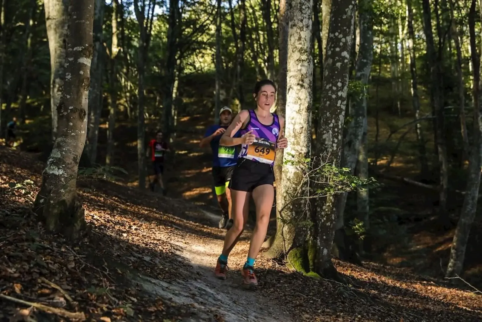 A female runner with bib number 649 is running on a wooded trail, with other runners behind her.