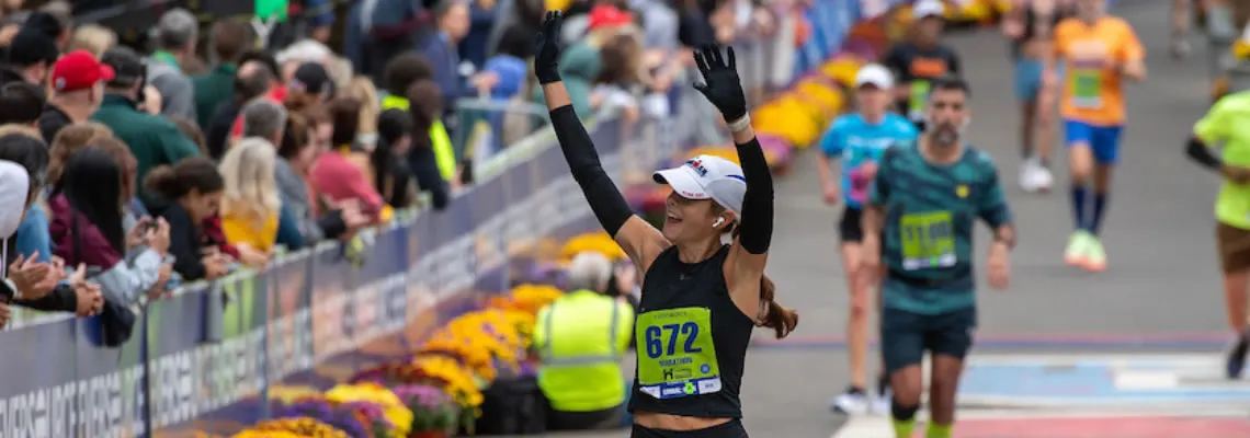 A female runner with her arms raised in celebration crosses the finish line, with a crowd cheering her on.