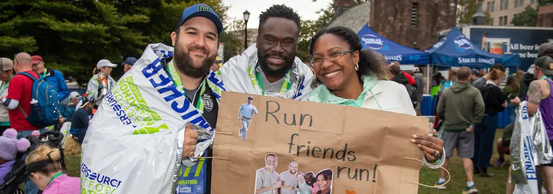 Three smiling runners, two wrapped in thermal blankets and one holding a handmade sign that says 'Run friends run!', pose for a photo at a race event.