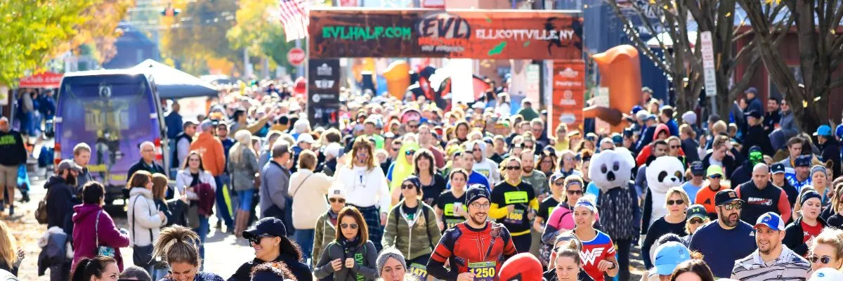 A large crowd of runners, some in costumes, gathered at the start line of the EVL Halloween Half Marathon under an archway.
