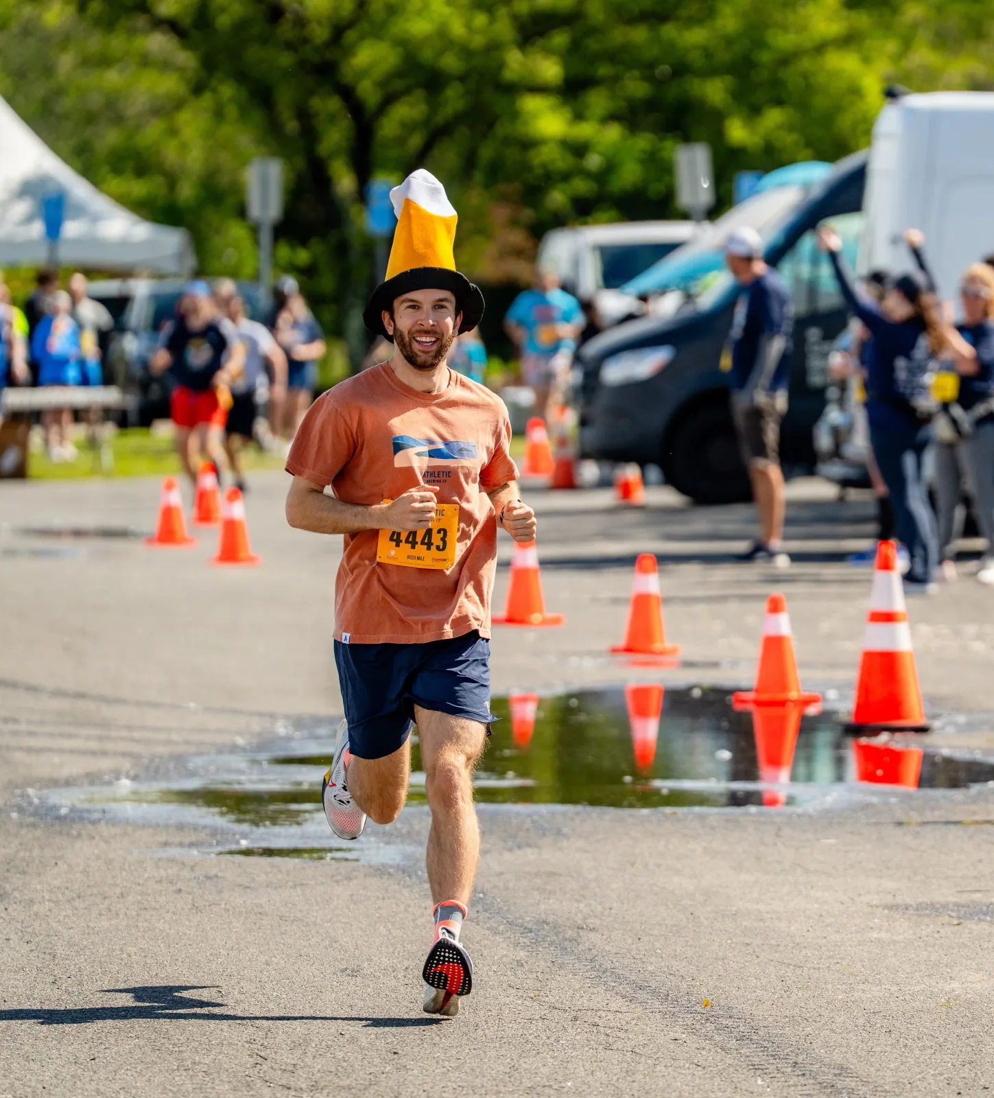 A smiling runner wearing a novelty beer-mug hat runs on a paved course, with orange cones and other runners in the background.