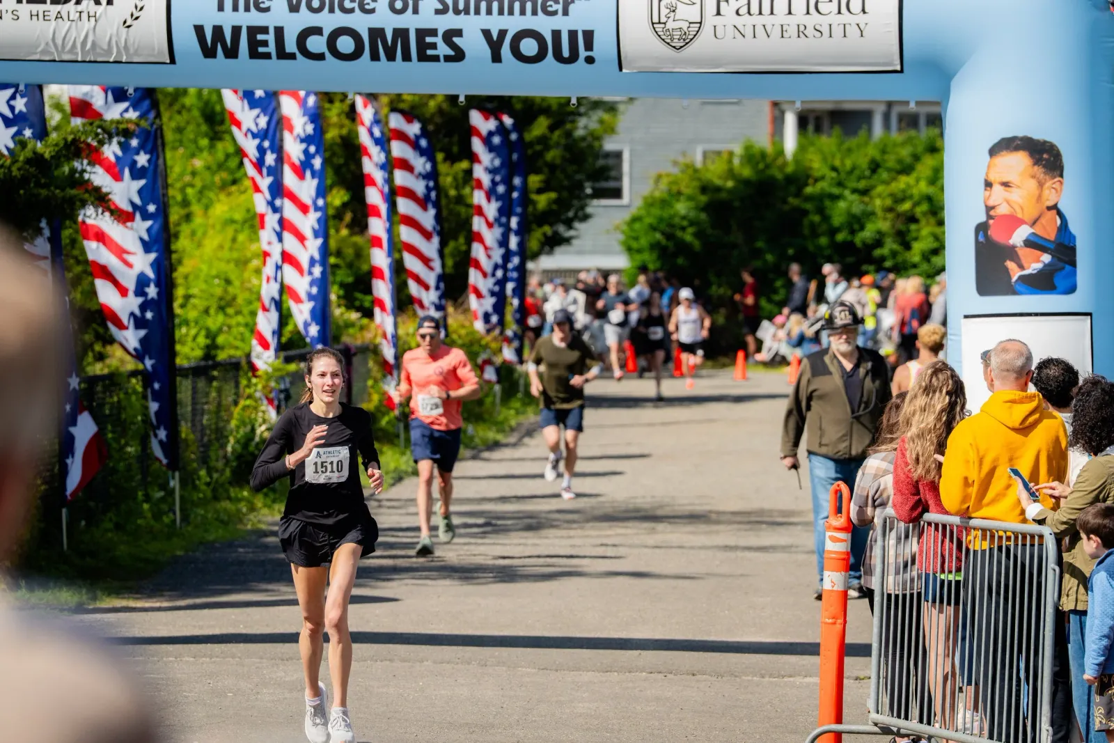 A female runner with bib number 1510 crosses the start line under a blue archway decorated with American flags.